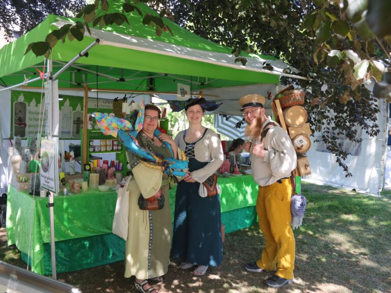 Persönliche Aufnahme von Angela Barbian am Marktstand der LH-Kerzenmanufaktur auf dem Flachsmarkt 2026 auf Burg Linn in Krefeld. In der Hand hält sie zwei dekorative Kerzen aus dem Sortiment der Manufaktur. Der Stand mit den farbenfrohen Kerzen und Duftkerzen vermittelt die kreative und handwerkliche Atmosphäre des traditionsreichen Kunsthandwerkermarktes.